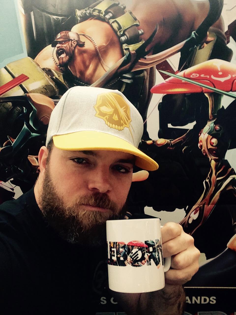 Devon in a Battleborn cap holding a Battleborn mug in front of character posters